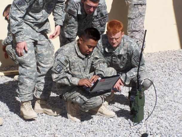 US Army soldier huddle over another soldier kneeling down with a military issue netbook