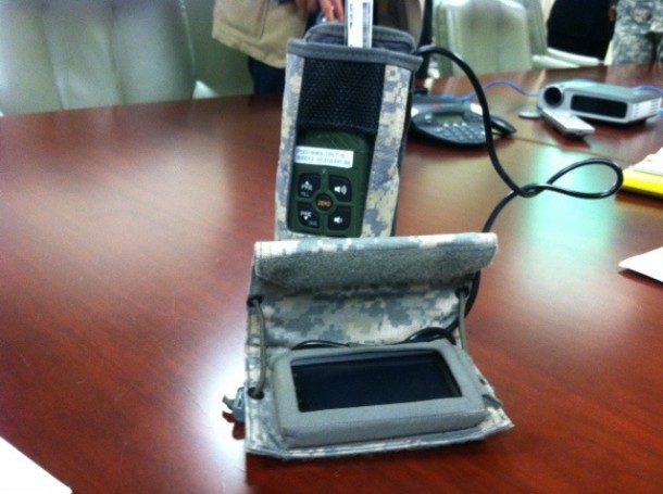 Smartphone with black cord connected to a military device. Both in camoflauge covering